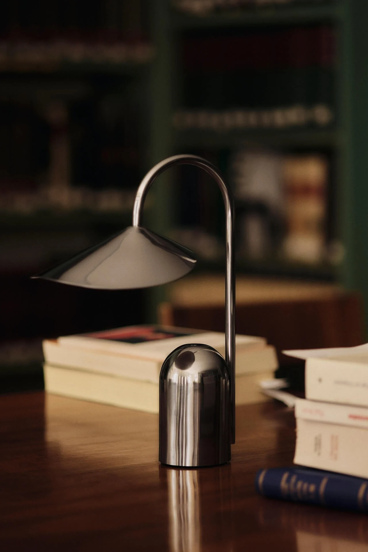 Een moderne zilveren tafellamp met een gebogen steel en conische kap staat op een houten bureau met vage boeken op de achtergrond.