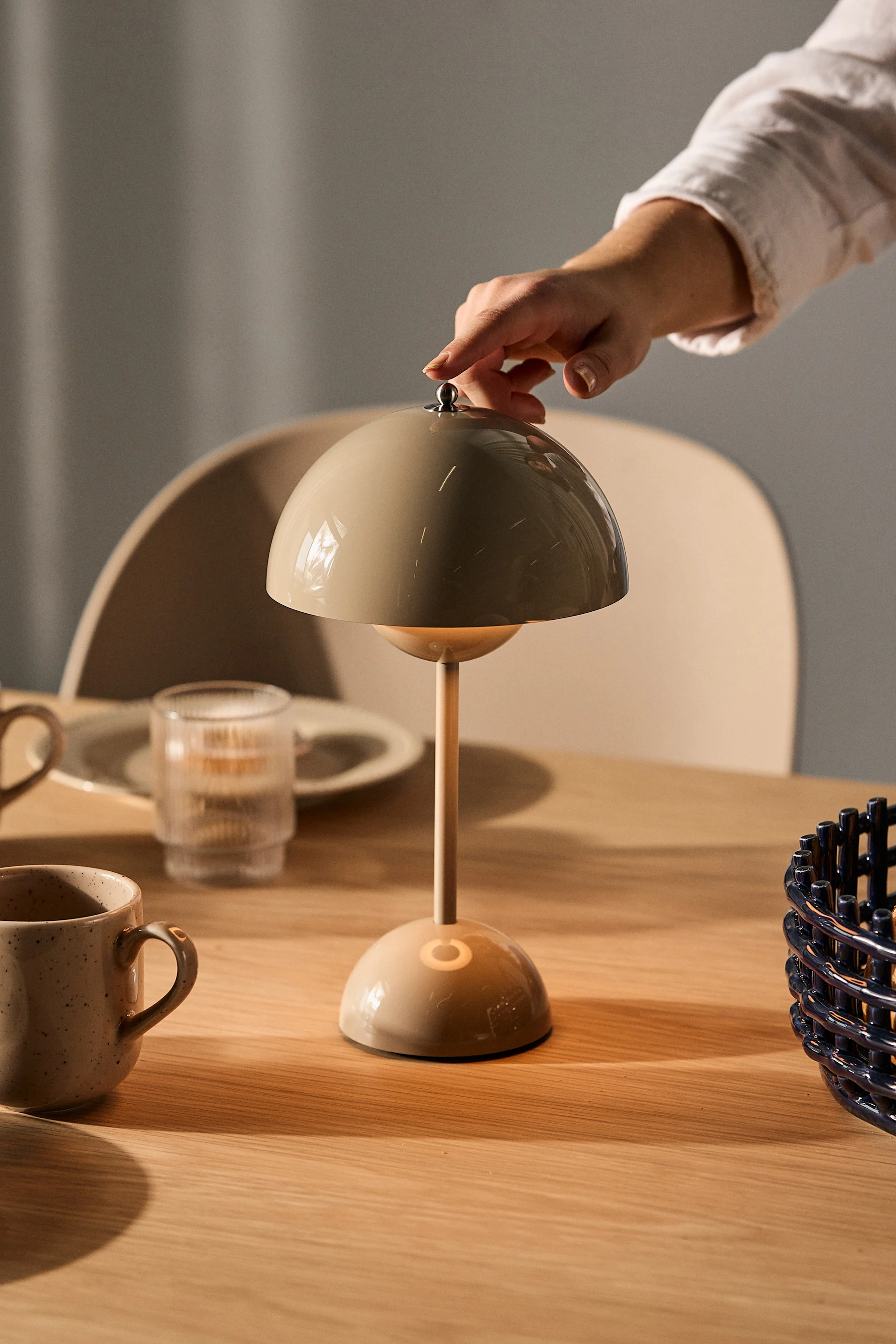 Een hand verstelt een beige paddestoelvormige tafellamp op een houten tafel met mokken en een glas.