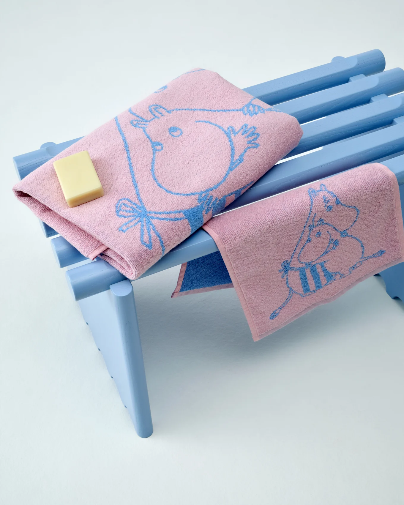 Twee roze Moomin handdoeken met blauwe patronen, één gevouwen met een stuk zeep, op een lichtblauwe bank.