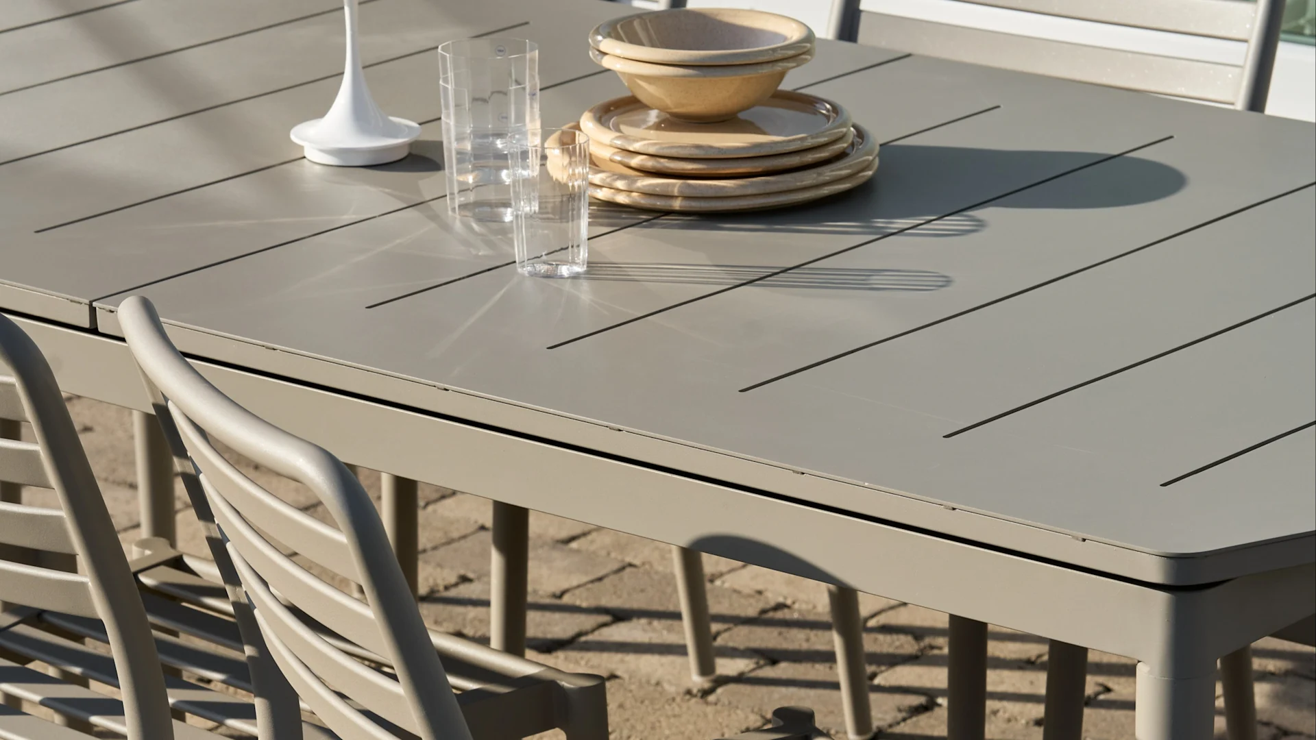 Lichtgrijze latten eettafel buiten met gestapelde beige borden en kommen, twee heldere glazen en een wit decoratief item.