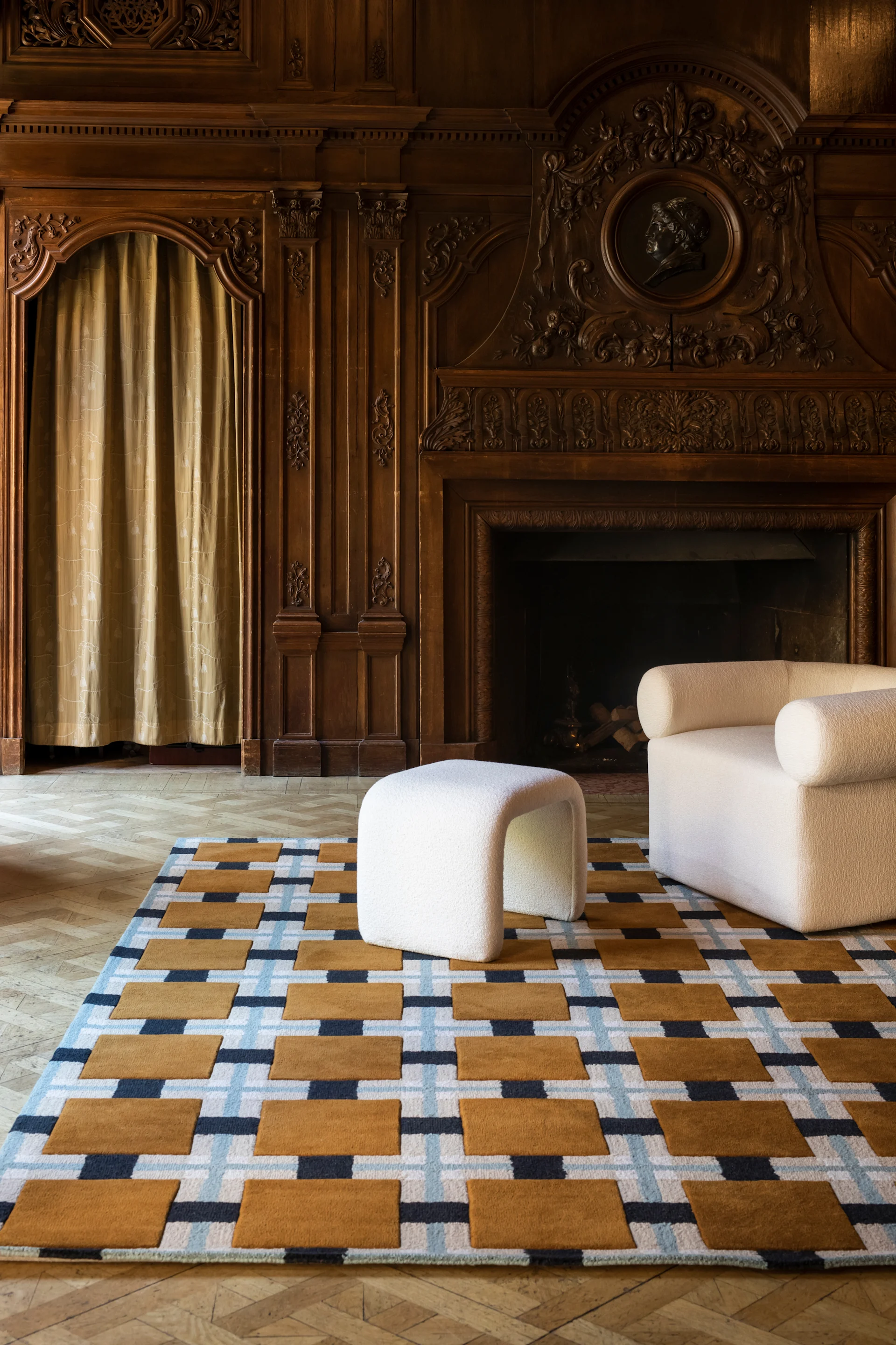 Geometrisch vloerkleed met bruine, blauwe, witte patronen, een witte getextureerde poef en fauteuil in een houten kamer.