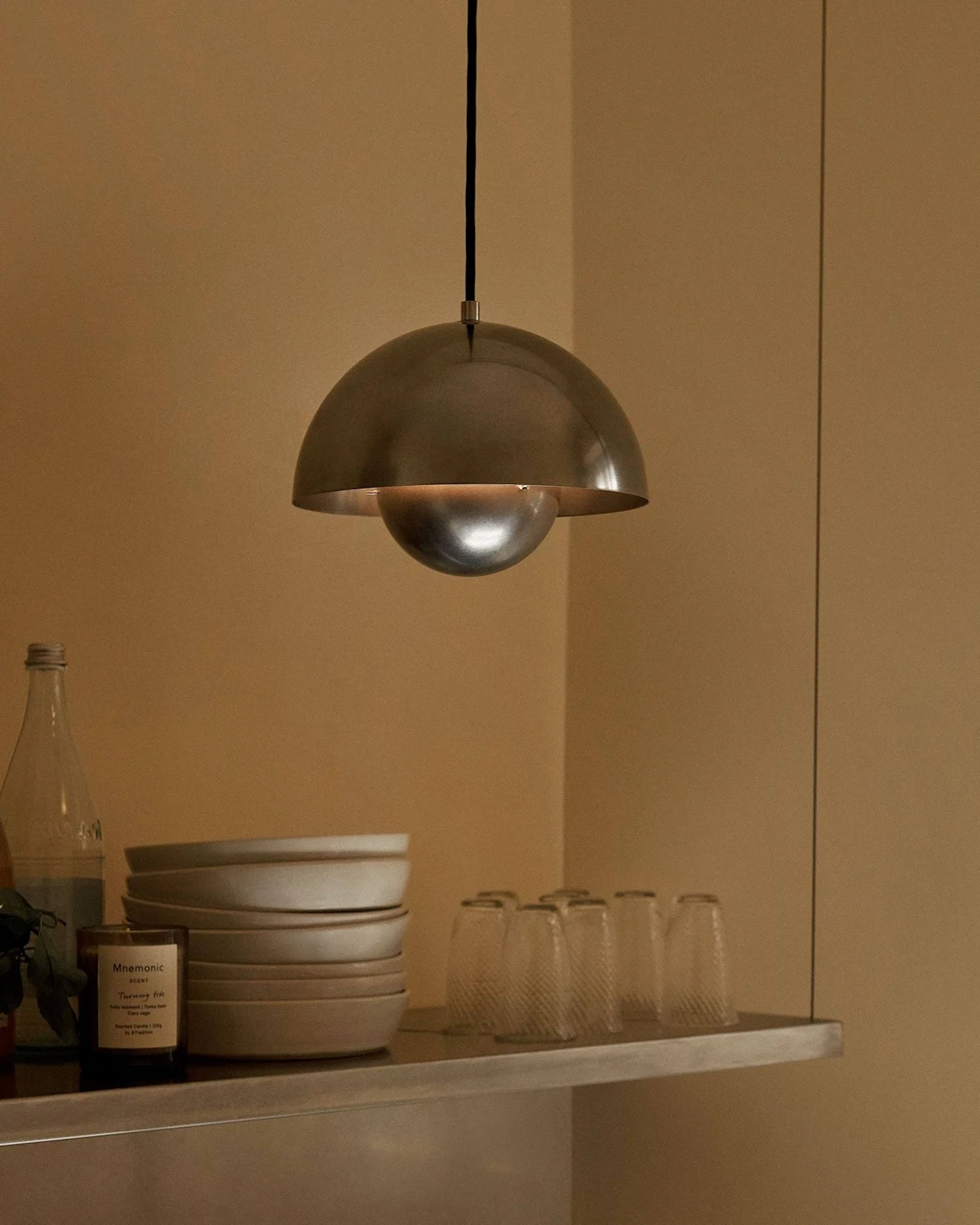 Een metalen koepelvormige hanglamp hangt boven een plank met kommen, glazen, flessen, een vrucht en een geurkaars.