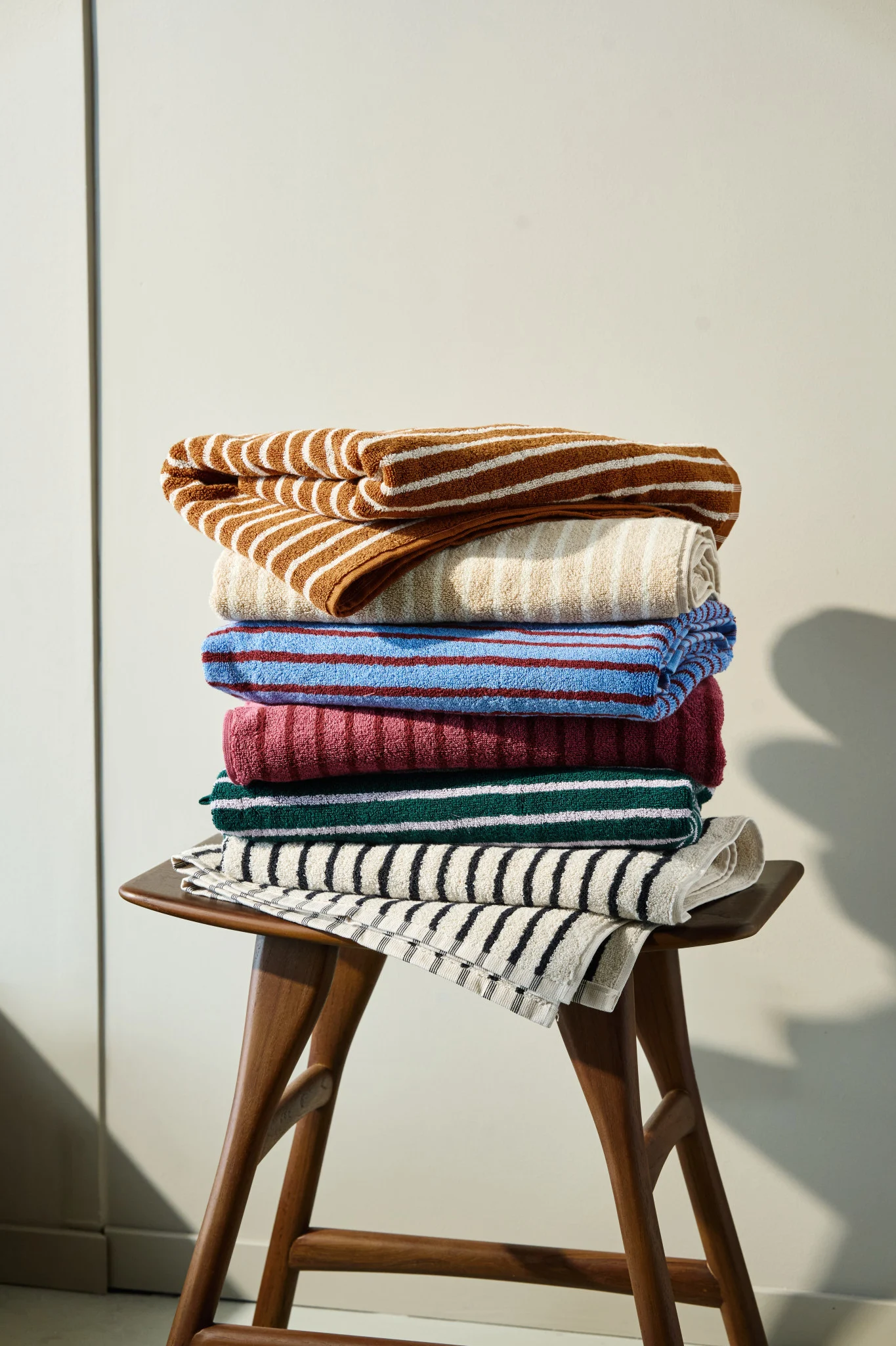 Een stapel kleurrijke gestreepte en effen handdoeken in bruin, beige, blauw, bordeaux, groen en zwart op een houten kruk.