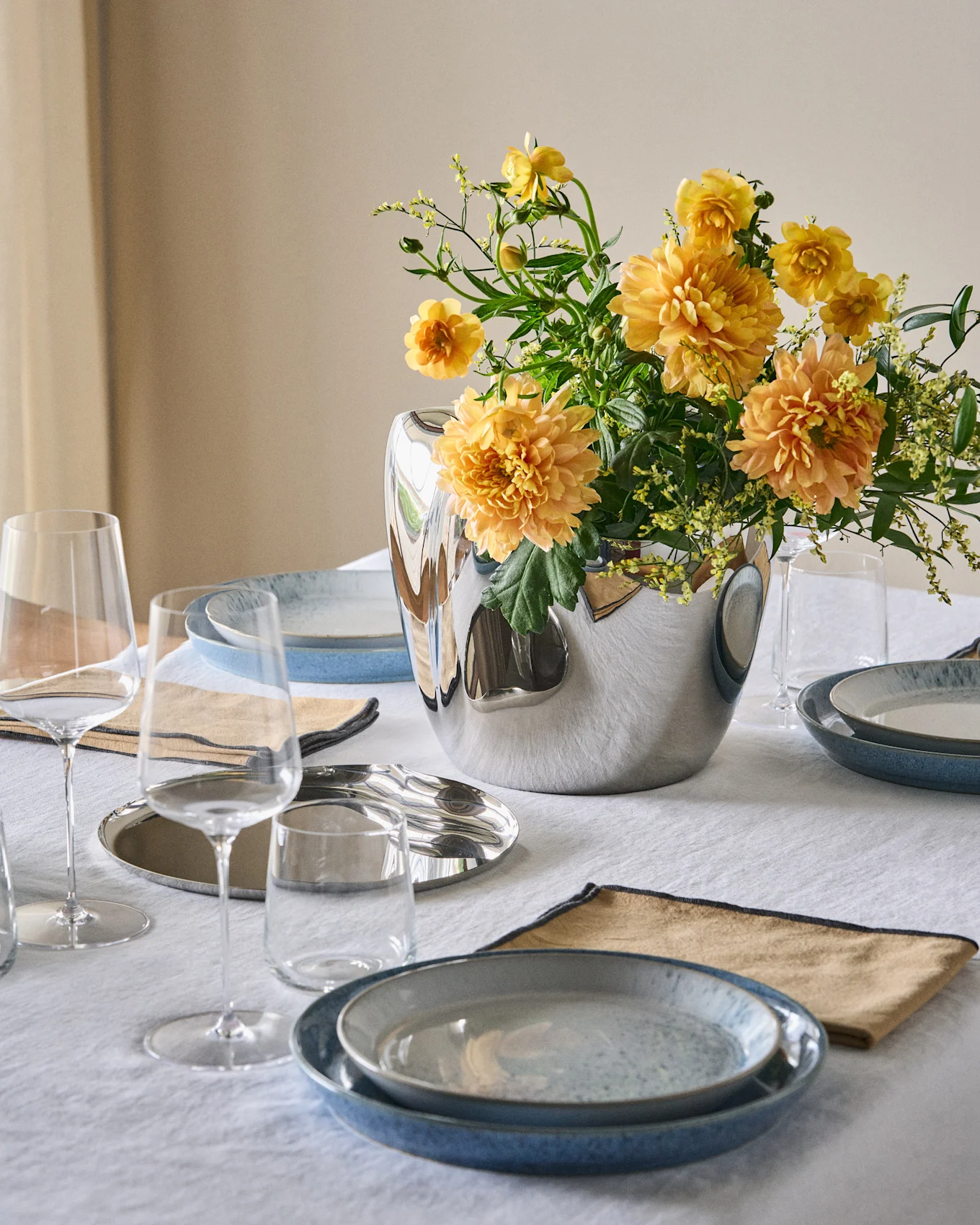Een eettafel gedekt met blauwe borden, beige servetten, heldere glazen en een zilveren vaas met oranje-gele bloemen op een wit tafelkleed.