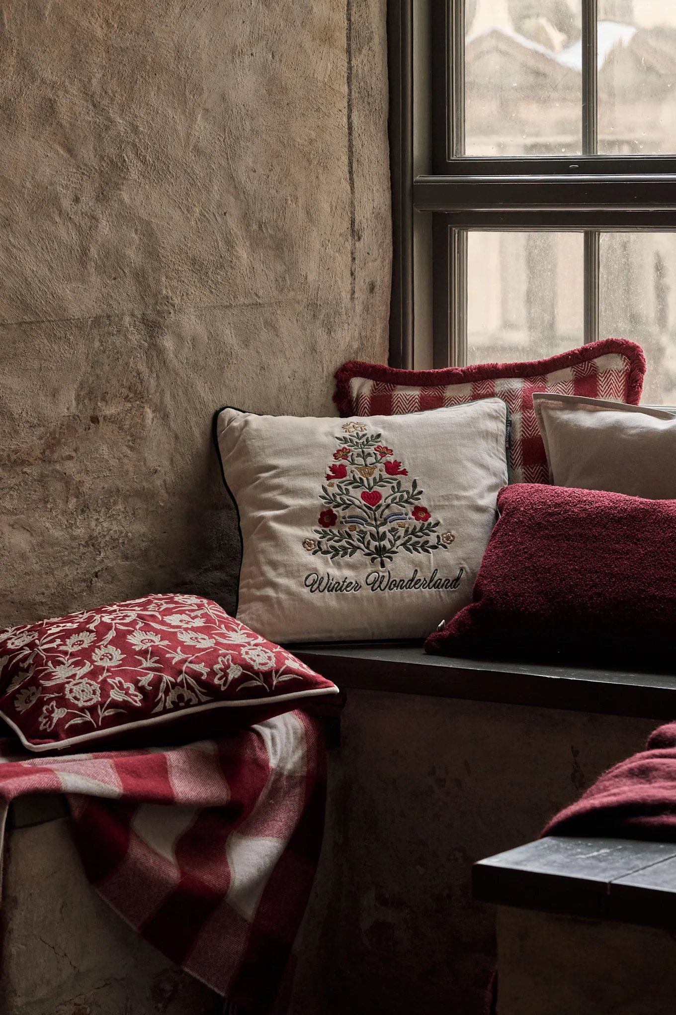 Gezellige vensternis met feestelijke kussens, waaronder een met een geborduurde kerstboom en 'Winter Wonderland', en een rood geruite deken.