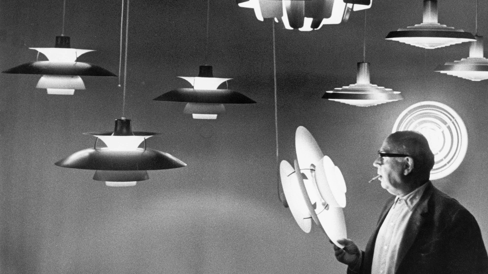 Zwart-wit foto van een man die een gelaagd lampdeel vasthoudt, omringd door vergelijkbare hanglampen.