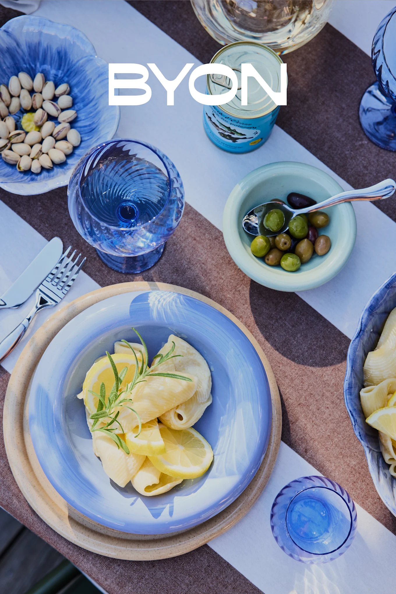 Bovenaanzicht van een eettafel met blauw servies, inclusief een bord pasta met citroen en rozemarijn, kommen met pistachenoten en olijven, en blauwe glazen. Het BYON-logo is over elkaar heen geplaatst.