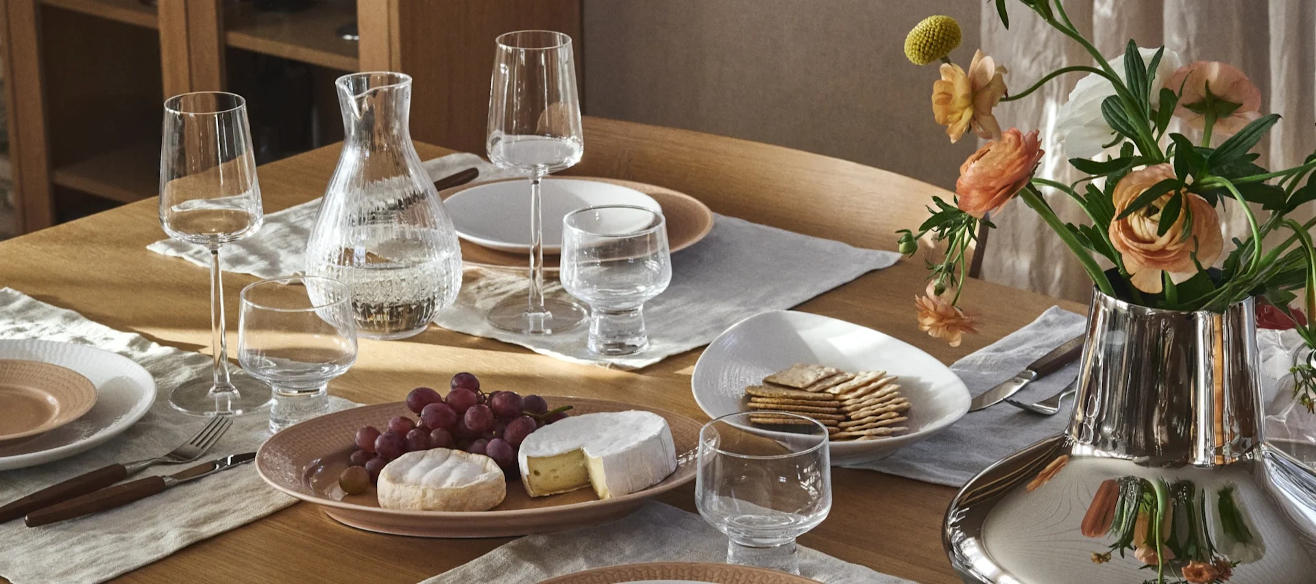 Een elegant gedekte eettafel met heldere glazen, een waterkan, lichtroze en witte borden, bestek, een kaas- en druivenplateau, crackers en een zilveren vaas met perzikkleurige bloemen.