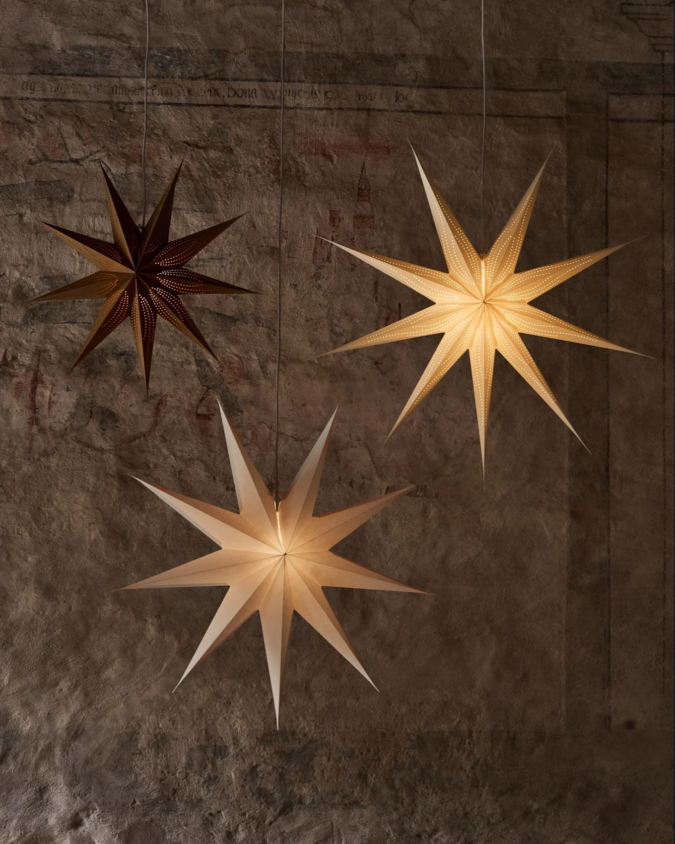 Drie stervormige papieren lantaarns hangen aan een getextureerde muur. Eén donkere lantaarn heeft kleine perforaties, twee lichtere verlichten de scène.