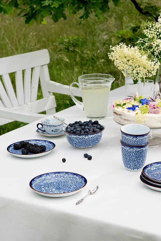 Buiten gedekte tafel met blauw-wit servies, verse bosbessen en bramen, limonadekan en een met bloemen versierde taart.