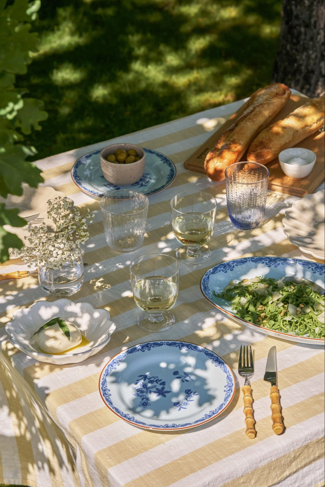 Een buitentafel gedekt met een geel-wit gestreept tafelkleed, burrata, salade, baguettes, olijven en blauw gedecoreerde borden.