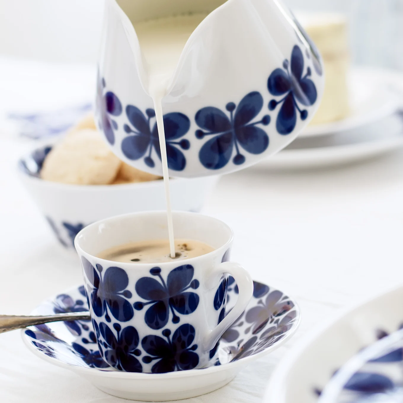 Melk wordt uit een blauw-wit bloemmotief melkkannetje in een bijpassende koffiekop op een schotel gegoten, met koekjes op de achtergrond.