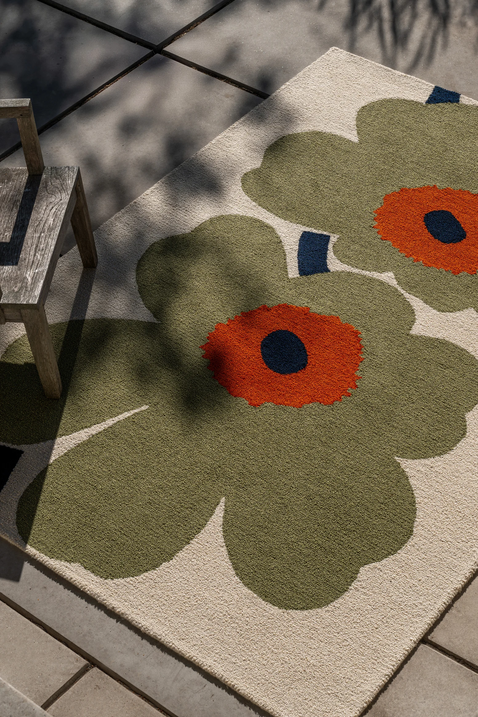 Vloerkleed met grote groene en oranje bloemmotieven op lichte achtergrond, onder houten stoel op terras, met bladschaduwen.
