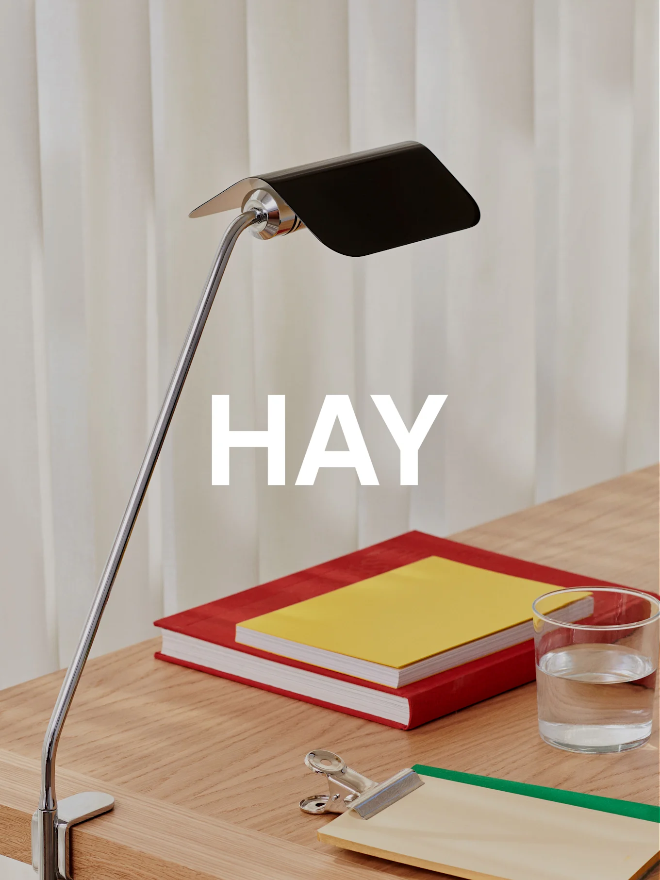 Een chromen bureaulamp met een zwarte kap verlicht een houten bureau met twee boeken, een glas water en een klembord.