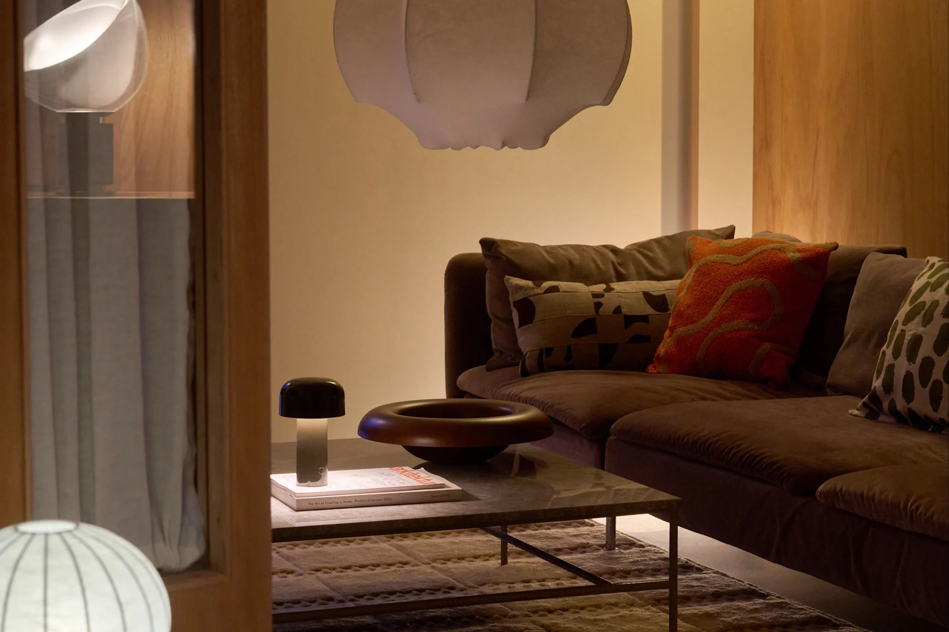 Knusse woonkamer met bruine bank, witte hanglamp, vloerlamp, salontafel, paddestoel lamp en decoratieve schaal.