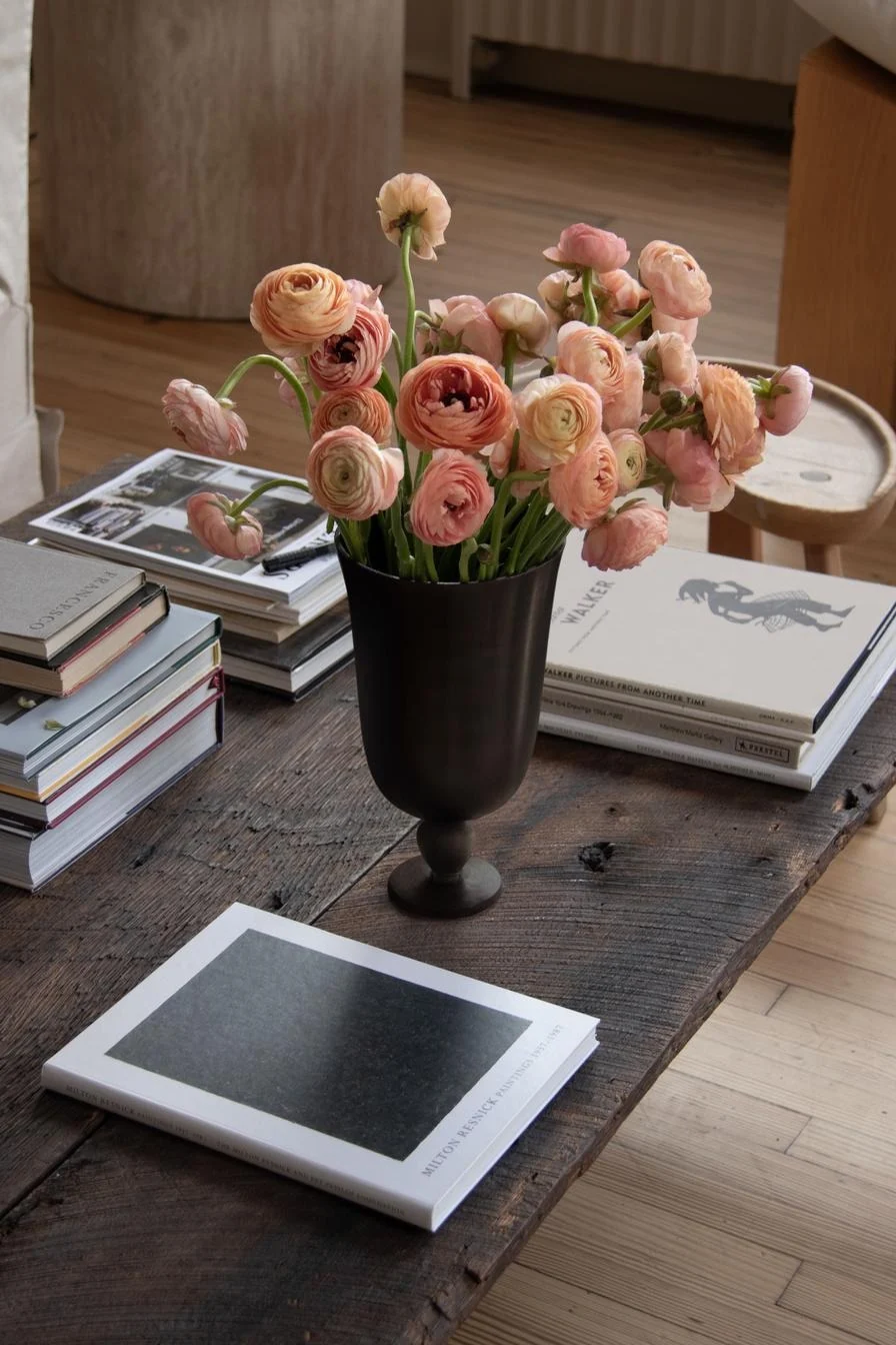 Perzikkleurige ranonkels in een donkere vaas op een houten salontafel omringd door kunstboeken.