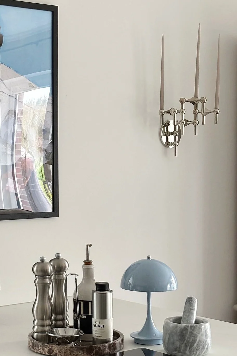 Een zilveren wandlamp met drie kaarsen hangt boven een witte tafel met keukenartikelen: molens, olieflesjes, een blauwe paddenstoellamp en een marmeren vijzel.