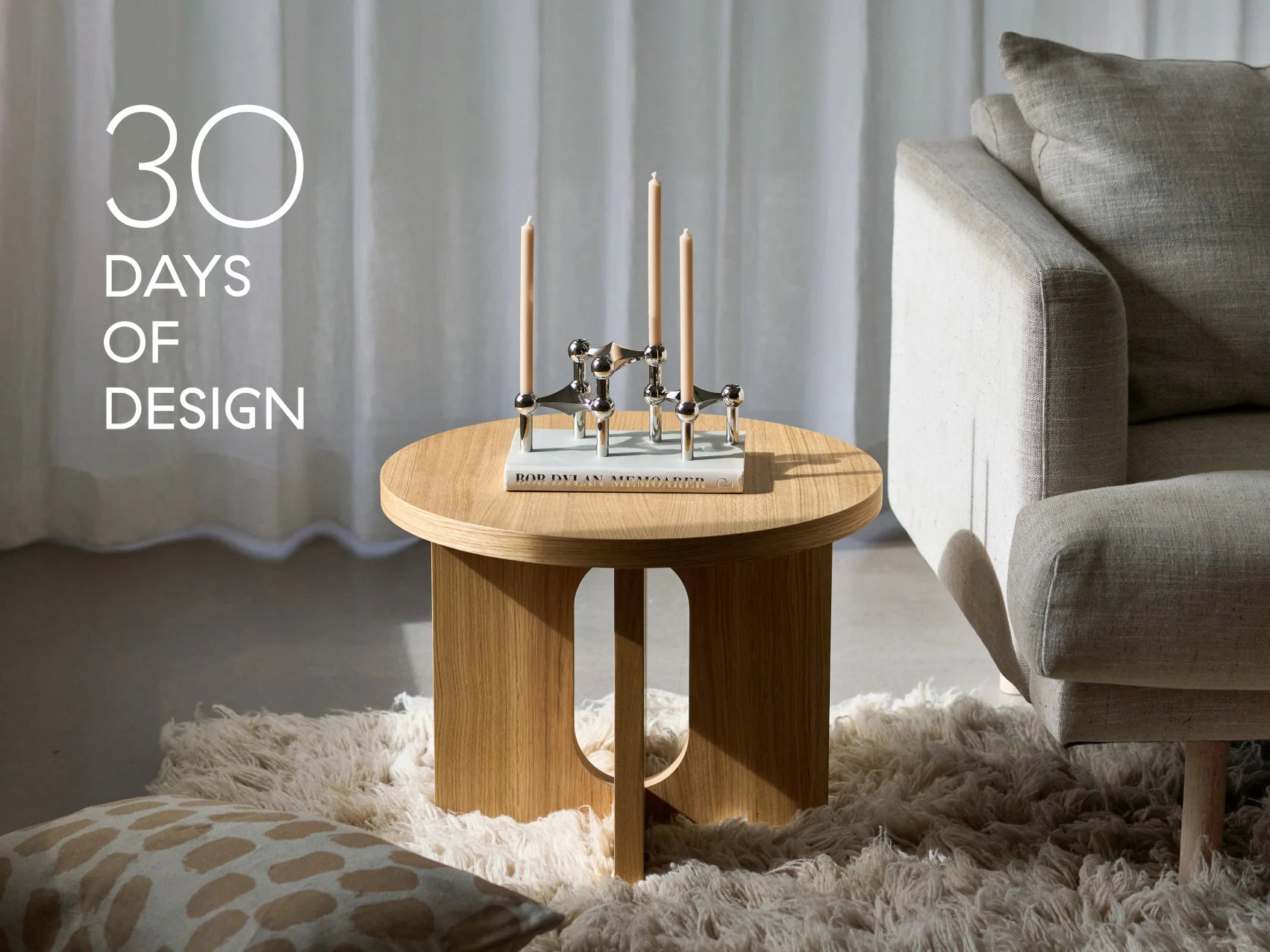 Moderne woonkamer met ronde houten bijzettafel, zilveren kandelaar met drie kaarsen, een boek en een beige bank. Tekst: '30 DAGEN DESIGN'.