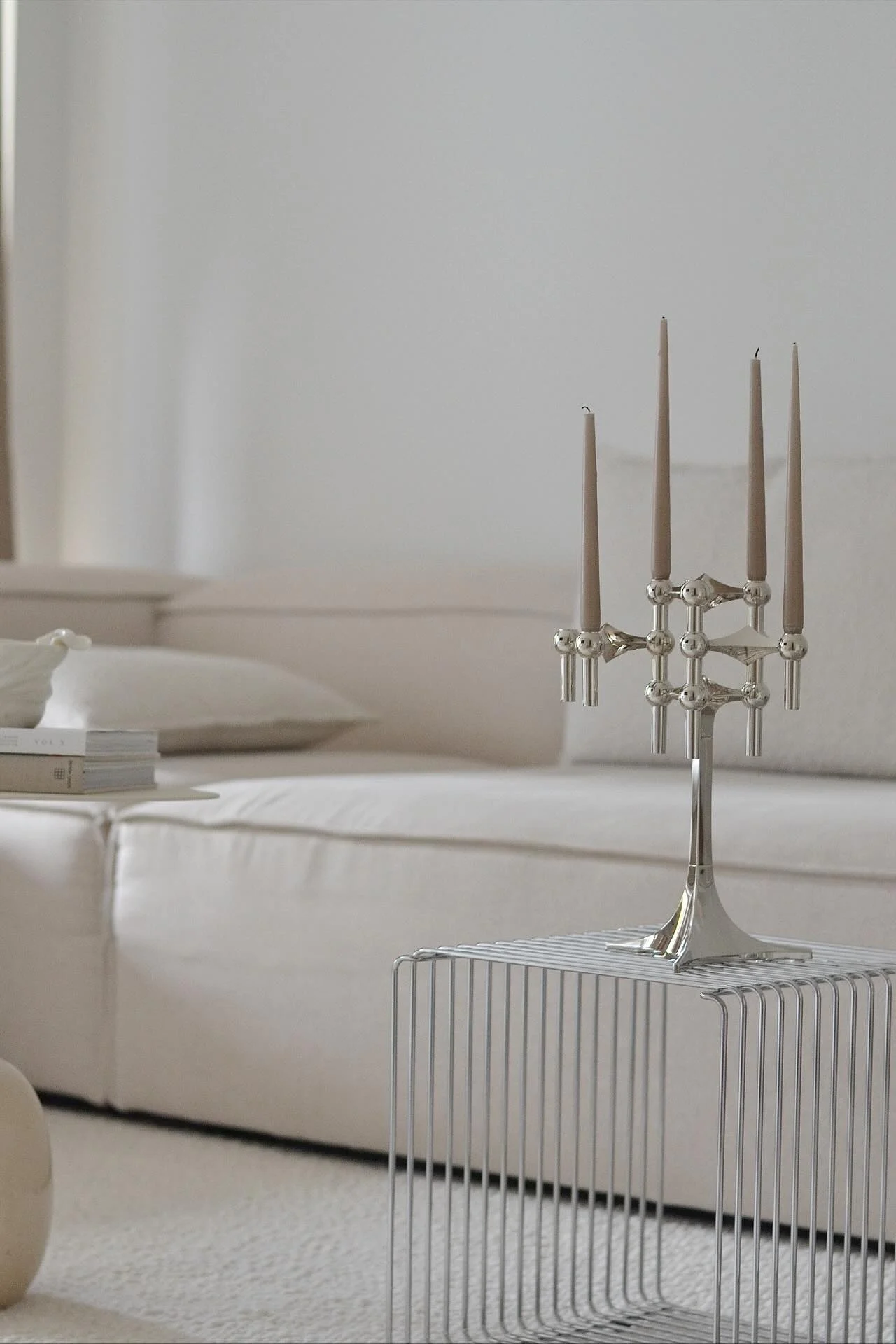 Een zilveren kandelaar met vijf lichtbruine kaarsen staat op een minimalistische metalen bijzettafel. Een lichte bank is op de achtergrond.