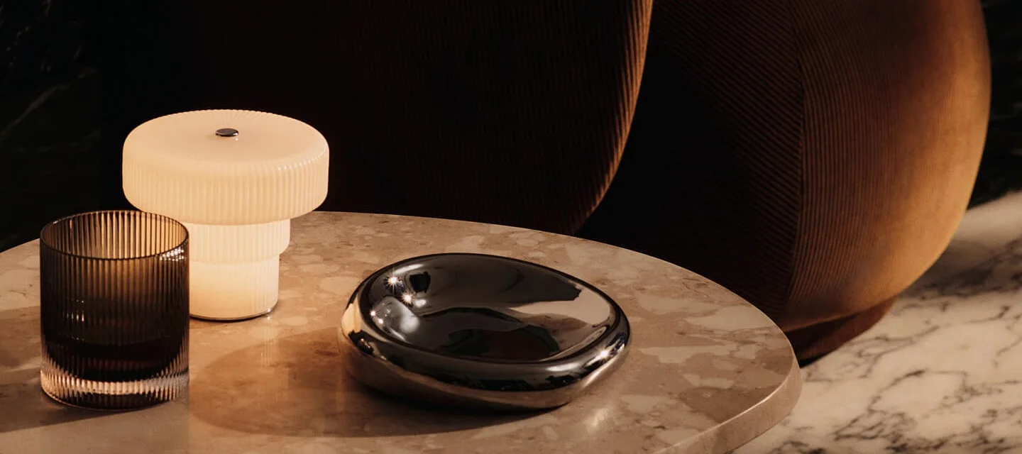 Een geribbelde tafellamp, donker glas en een glanzende decoratieve schaal op een marmeren tafel.