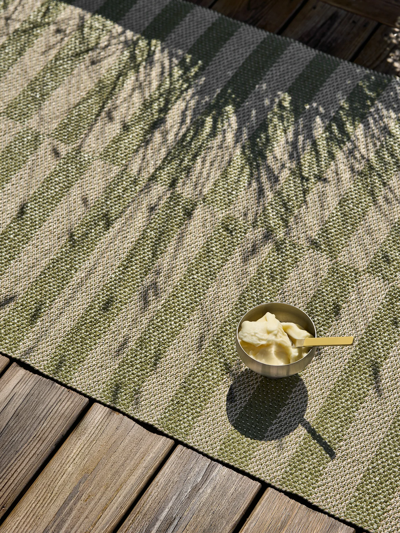 Een zilveren kom met dessert en een gele lepel staat op een groen-beige gestreepte buitenmat op een houten terras, besprenkeld met palmboomschaduwen.
