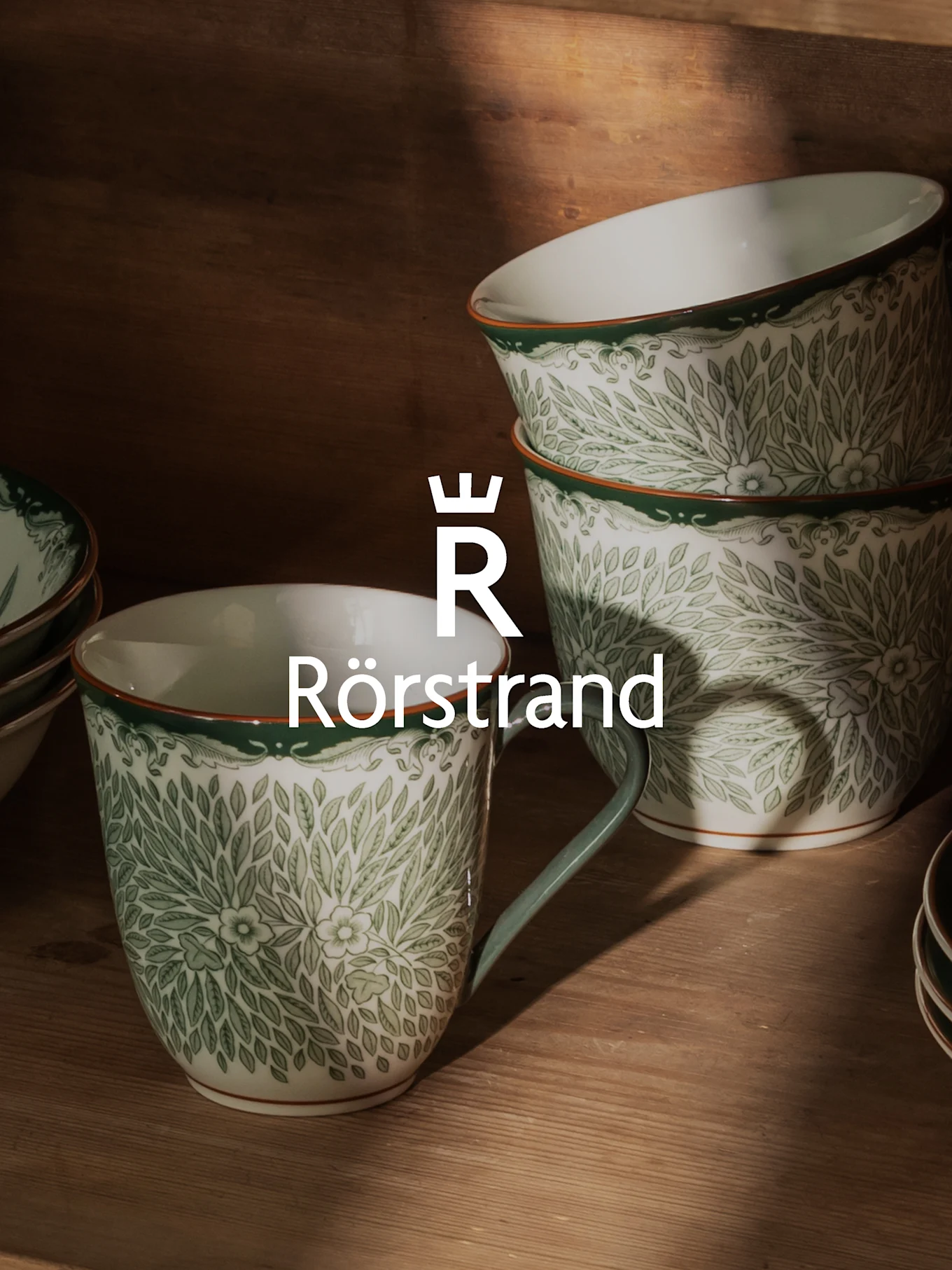 Witte keramische mokken en kommen met groen bladpatroon, oranje rand en Rörstrand-logo.