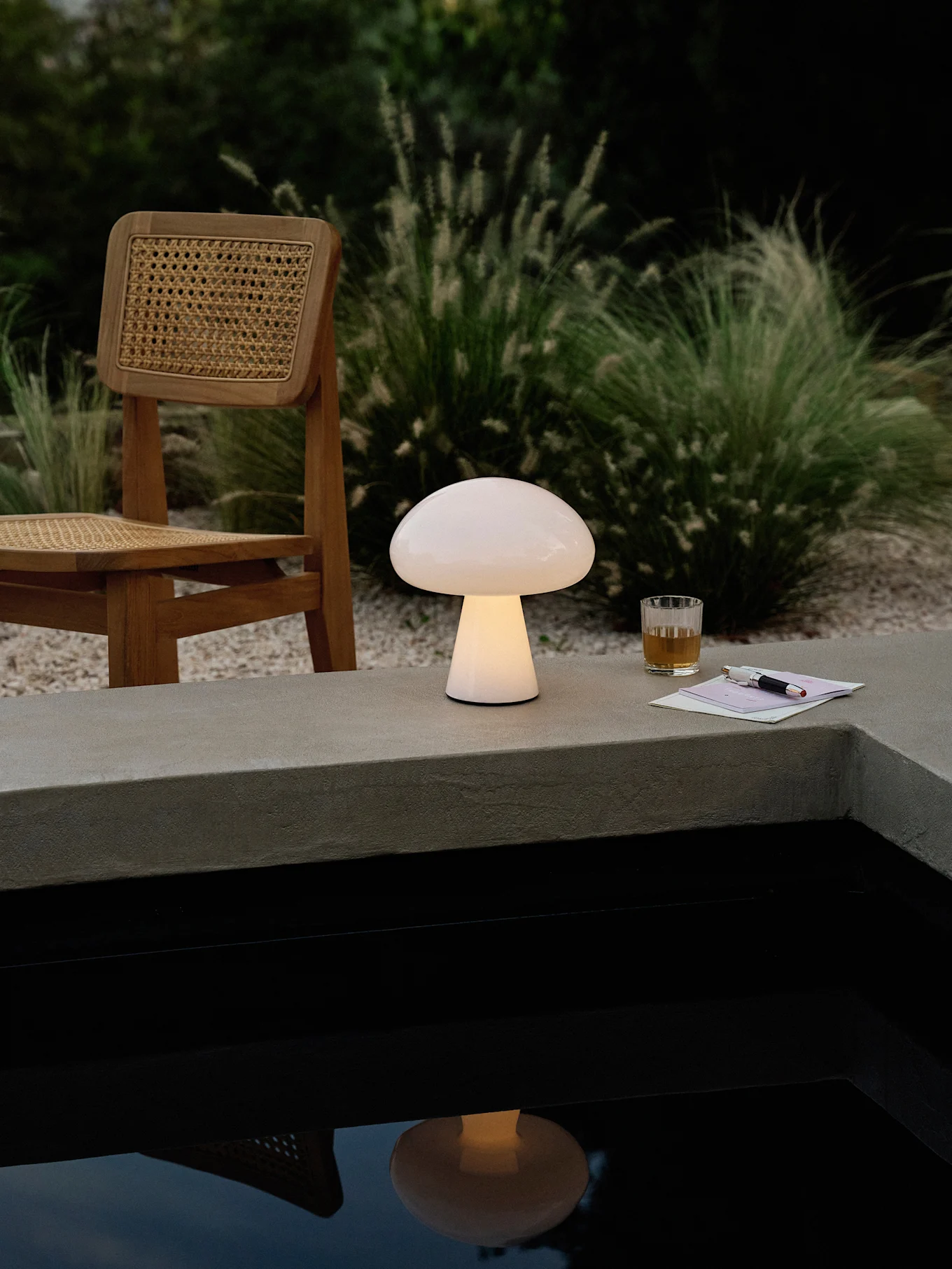 Een gloeiende paddestoelvormige lamp op een betonnen richel naast een zwembad, met een houten stoel, drankje en pen.