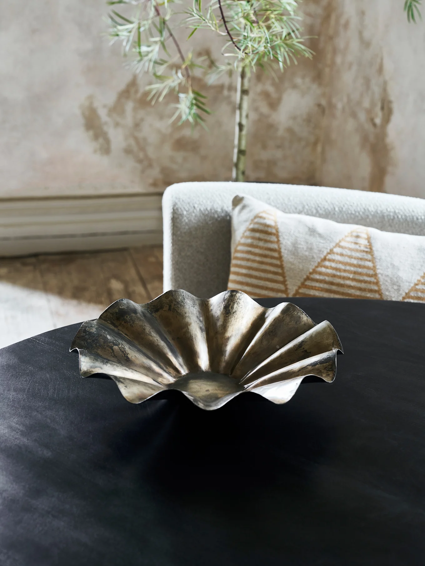 Een decoratieve metalen schaal met een golvende, gerimpelde rand staat op een donkere tafel in een woonkamer.