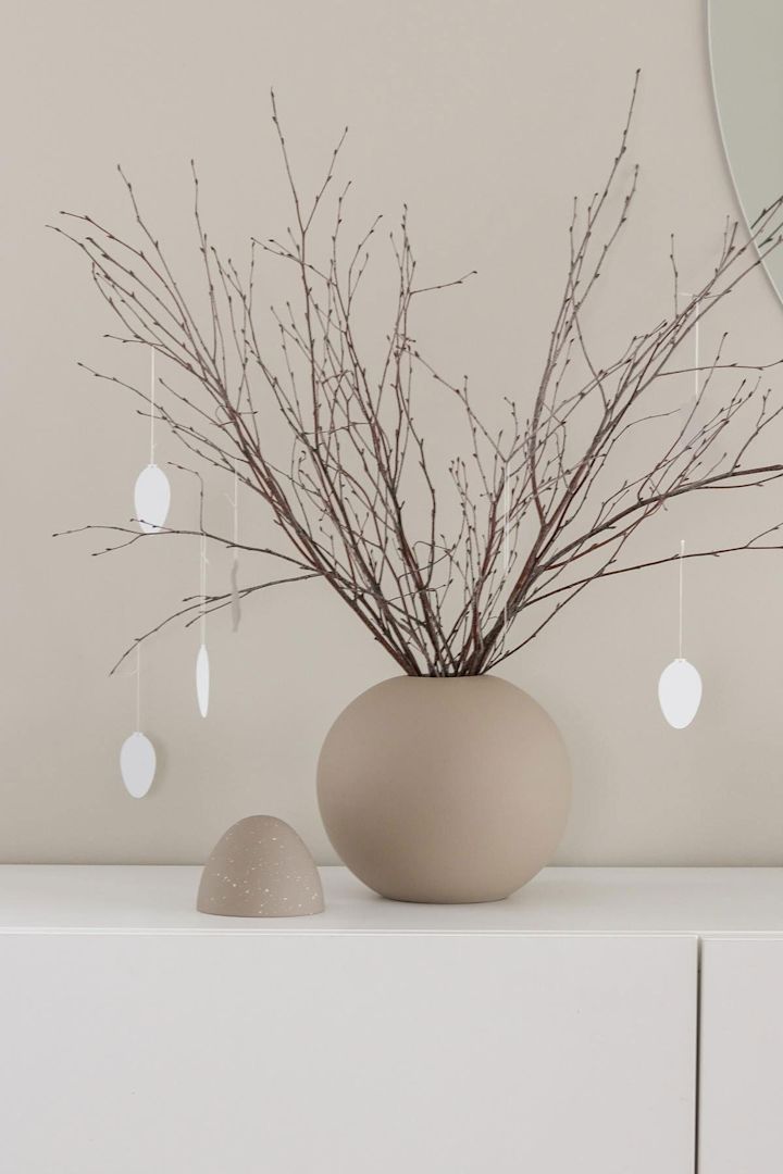 Een paastak staat ook op een wit dressoir in een zandkleurige bollenvaas met paasdeco-ei hangers, beide van Cooee Design, in het huis van @tunadalen.