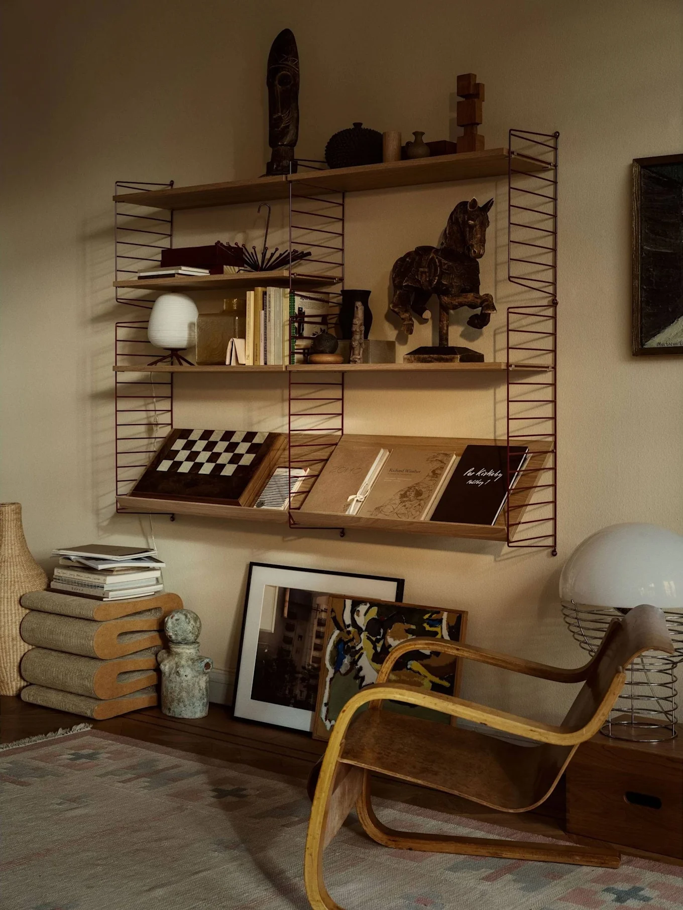 Een gezellige kamer met een wandrek gevuld met boeken, kunst en decoratie, naast een gebogen houten fauteuil en een grote witte lamp.