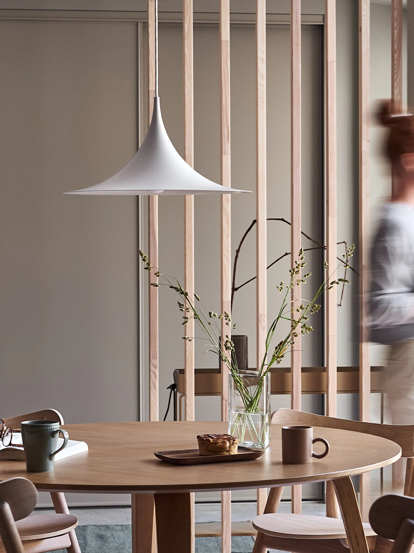 Moderne eethoek met ronde houten tafel, witte pendellamp, vaas met grassen, gebakje en mokken, met een vaag persoon die voorbij loopt.