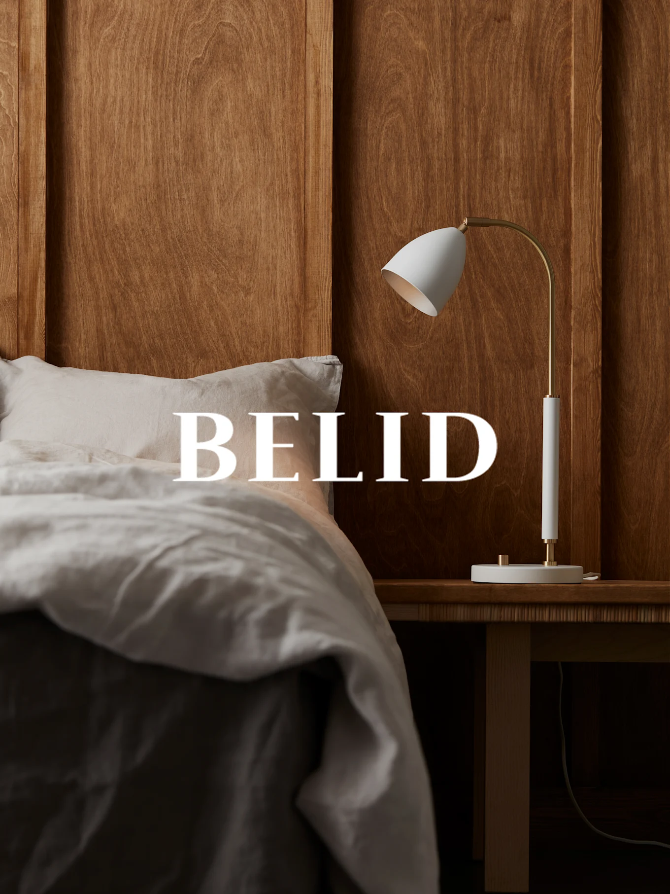 Witte en messing lamp op een houten nachtkastje, met 'BELID' tekst boven een bed met linnen lakens.