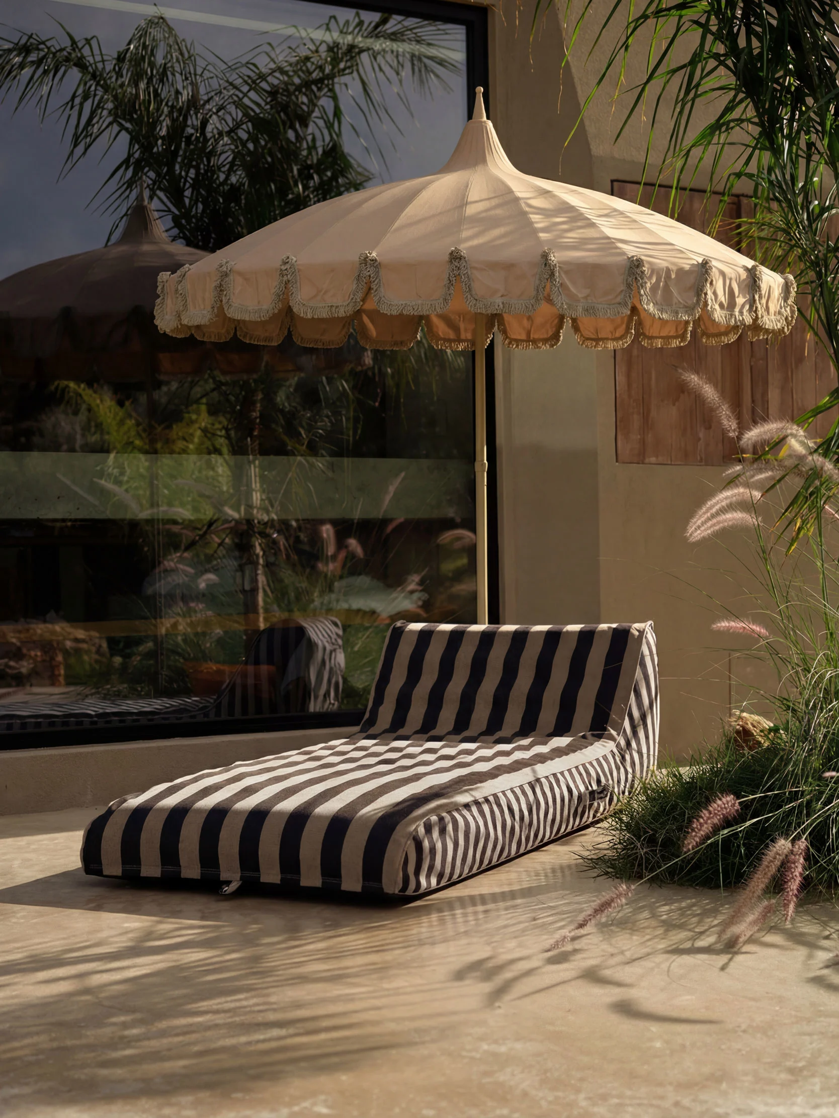 Buitenpatio met een beige parasol met franjes en een gestreept loungekussen op lichte vloer, omringd door planten.
