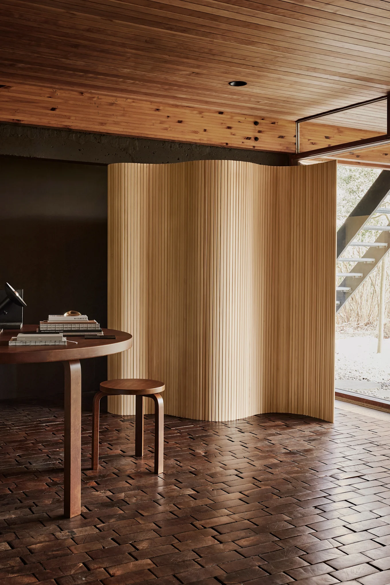 Moderne kamer met een ronde houten tafel, een kleine kruk en een golvend houten scherm. Donkere houten vloer, licht houten lattenplafond en een raam naar een buitentrap.