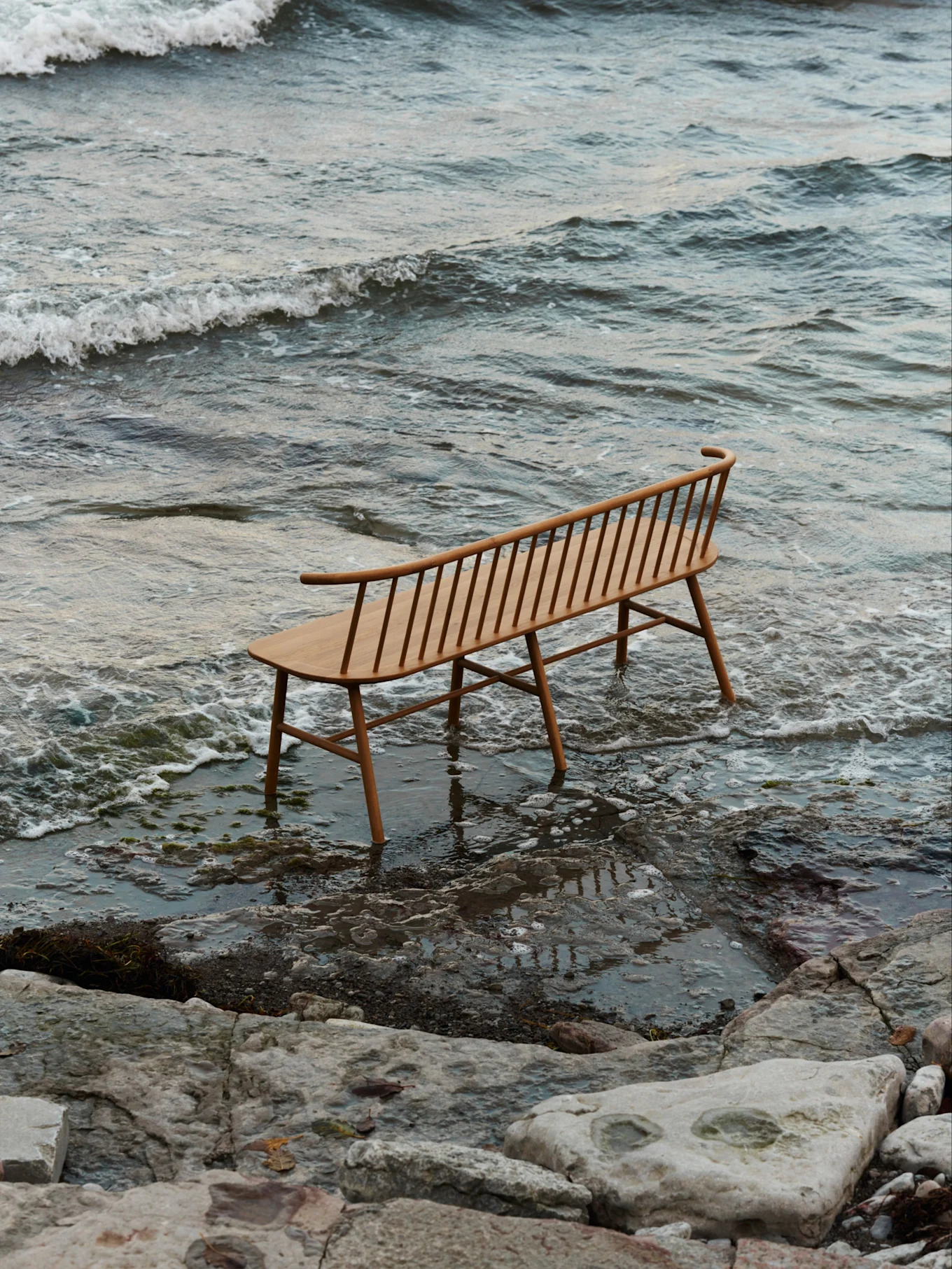 Een lichte houten lattenbank met een gebogen rugleuning staat in ondiep water op een rotsachtig strand met zachte golven.