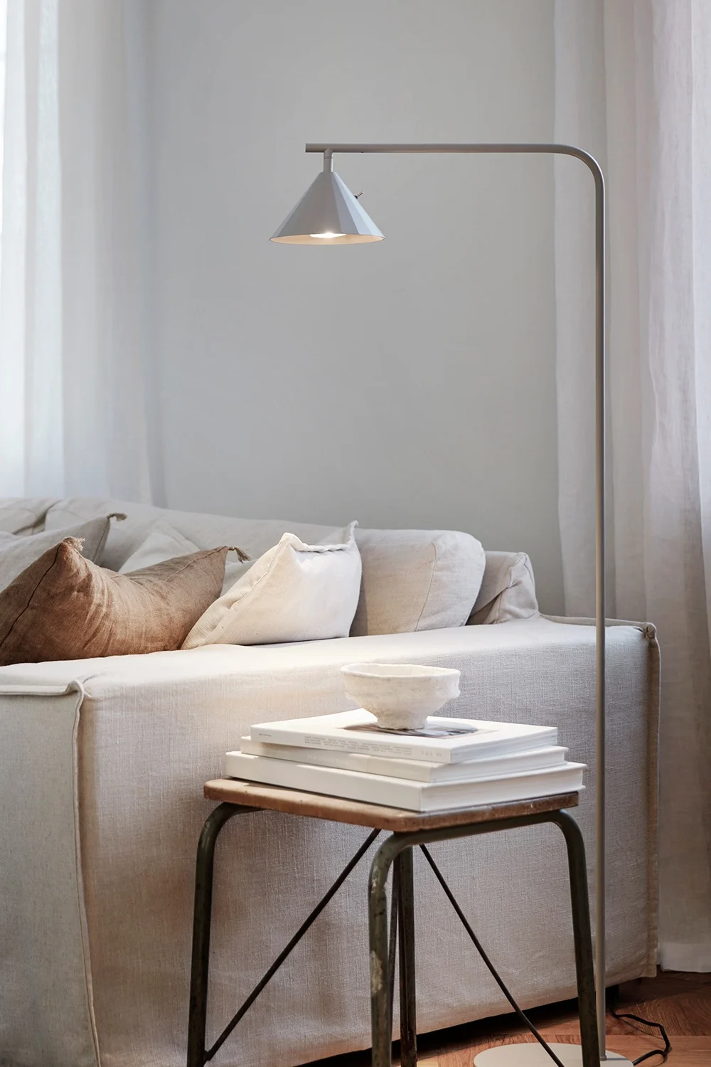 Een gezellige woonkamer met een lichtgekleurde linnenbank, vloerlamp en bijzettafel met boeken en kom.
