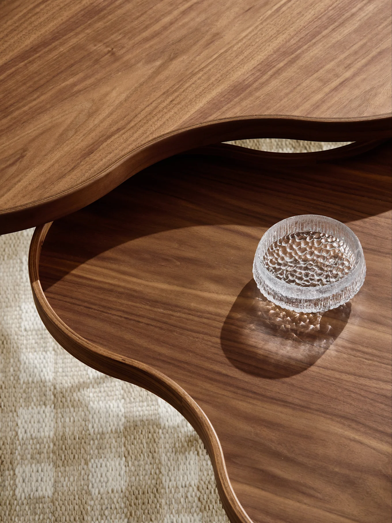 Close-up van een meerlagige houten salontafel met golvende randen en getextureerde glazen kom op onderste laag, op getextureerd kleed.