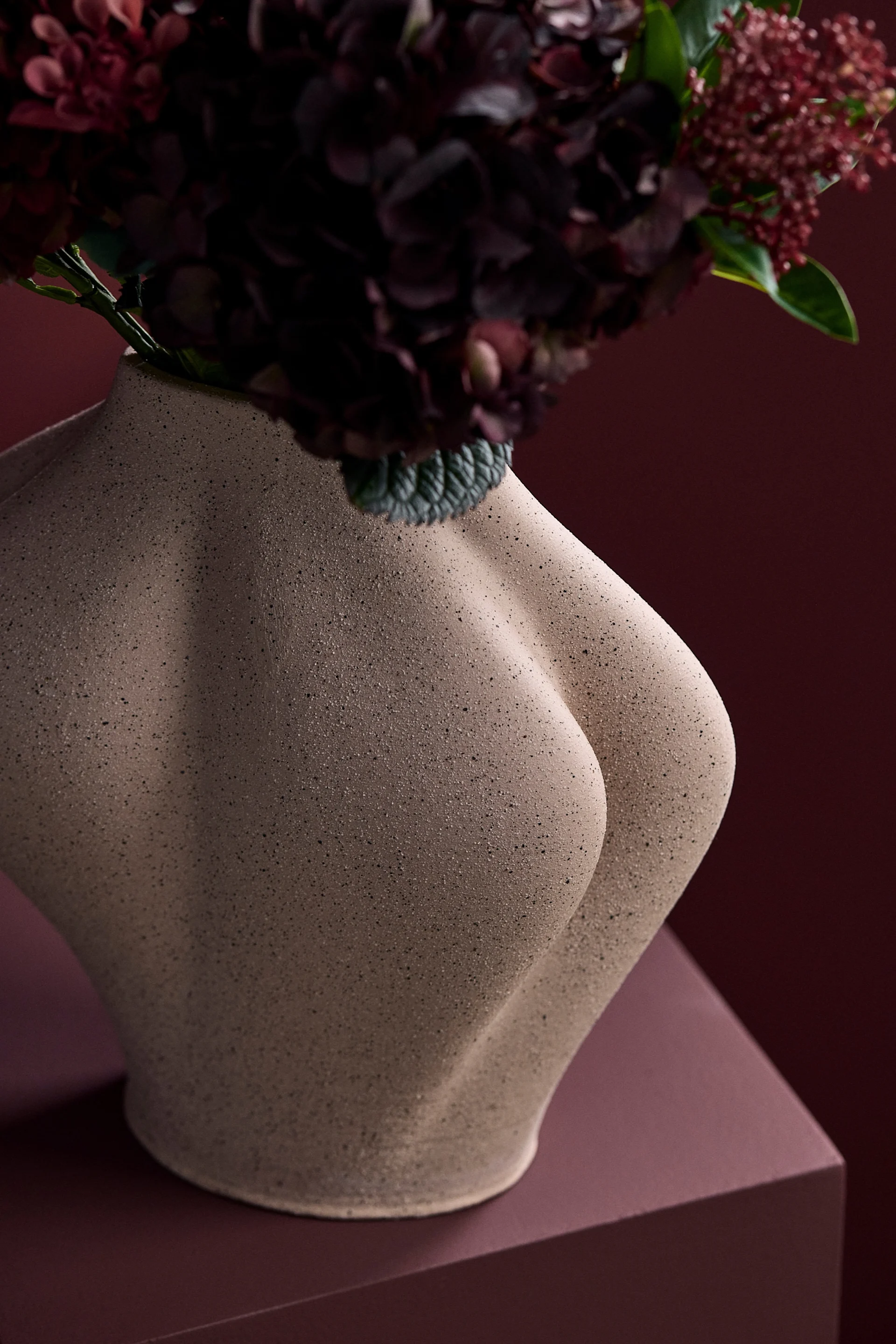 Sculpturale beige vaas in de vorm van een menselijke torso, met donkerrode bloemen tegen een bijpassende rode achtergrond.