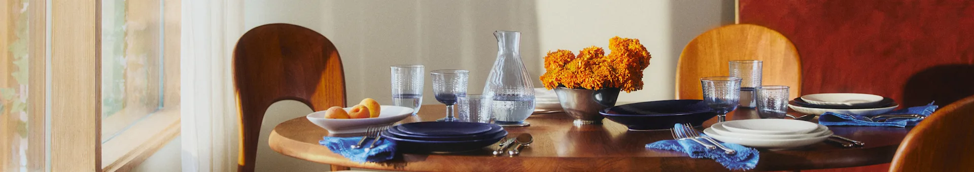 Ronde houten eettafel met blauw-witte borden, blauwe servetten en oranje bloemen. Zonlicht filtert door een raam naar binnen.