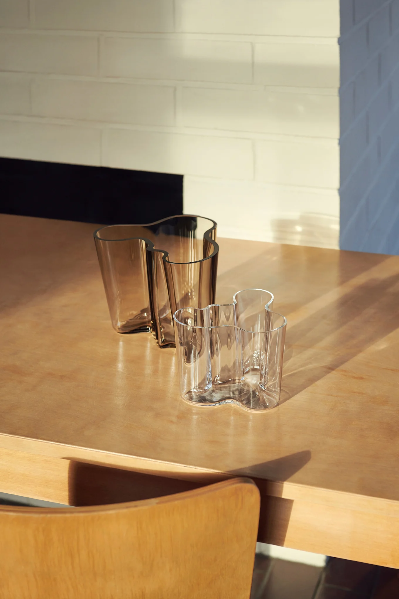Drie golvende glazen vazen op een houten tafel. Twee zijn amberkleurig, één is helder, met zonlicht en schaduwen over de scène.