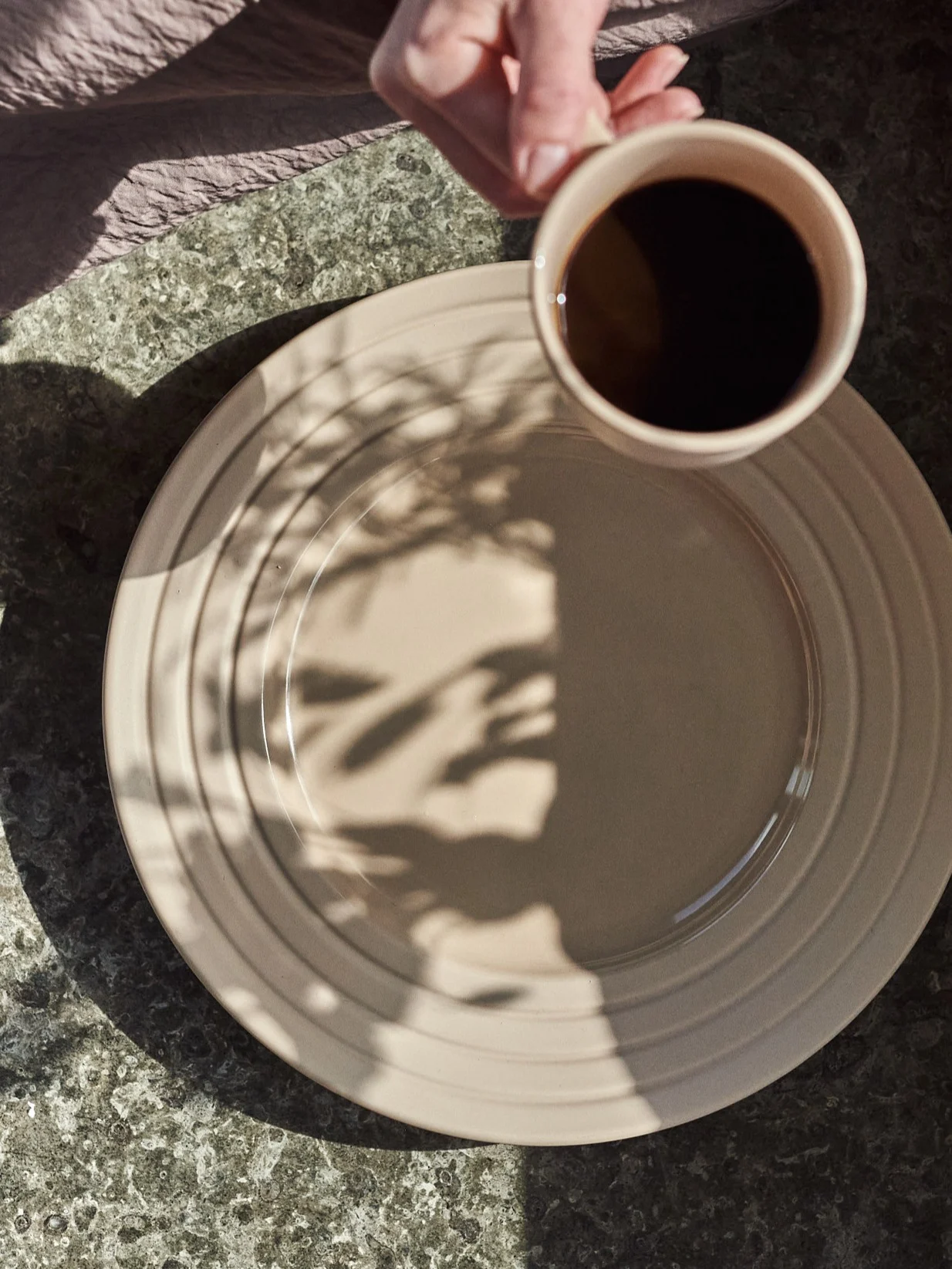 Hand houdt koffiekop boven beige bord met bladschaduwen op een gespikkelde tafel.