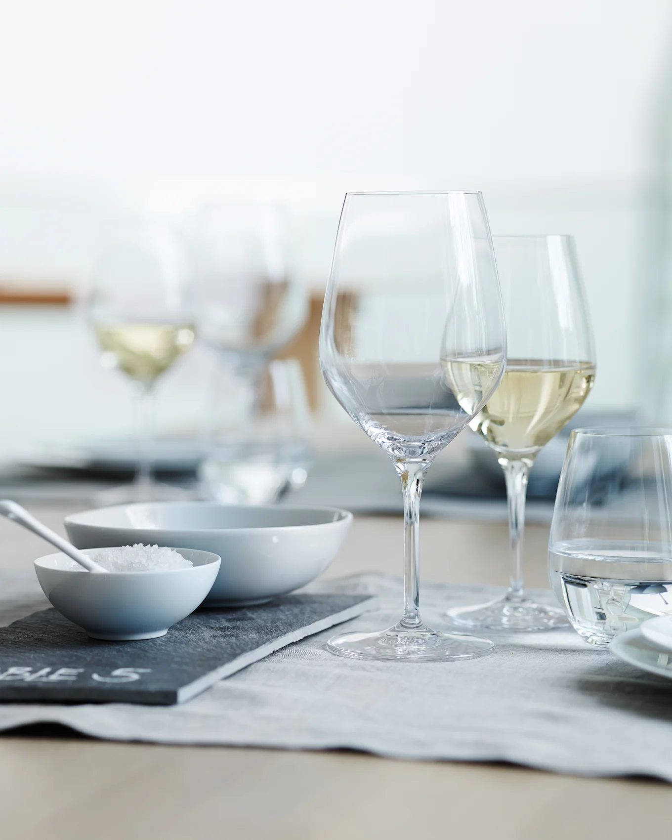 Een gedekte eettafel met heldere wijn- en waterglazen, een kom met grof zout en een lege kom op een grijze placemat.