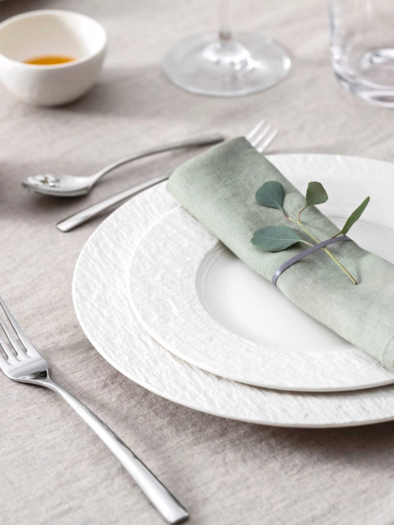 Elegante tafelschikking met lichtgroen linnen servet, eucalyptusbladeren, witte gestructureerde borden en bestek op een beige tafelkleed.