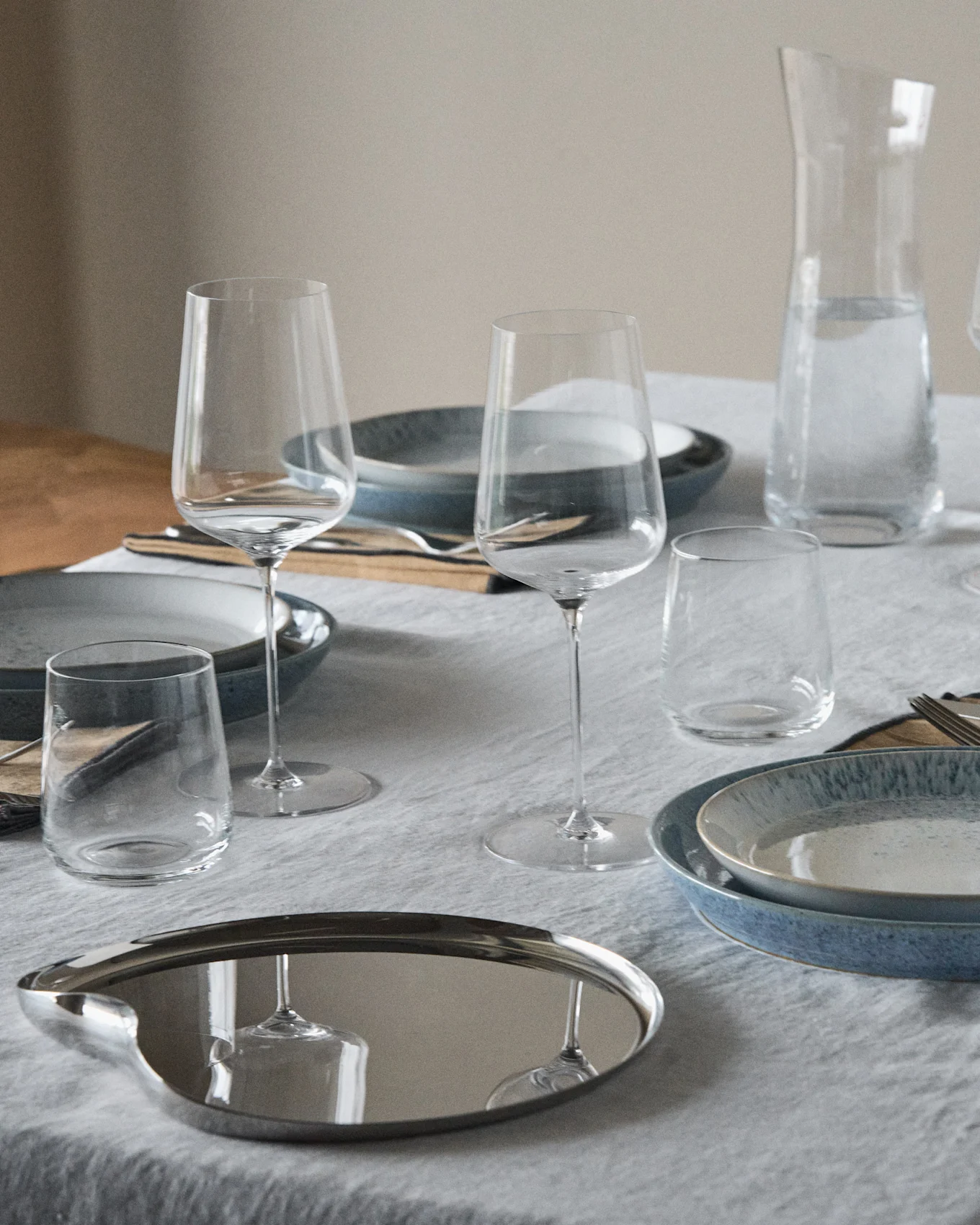 Een gedekte tafel met blauwe en gespikkelde borden, wijnglazen, glazen, een waterkan en een reflecterend ovaal dienblad.