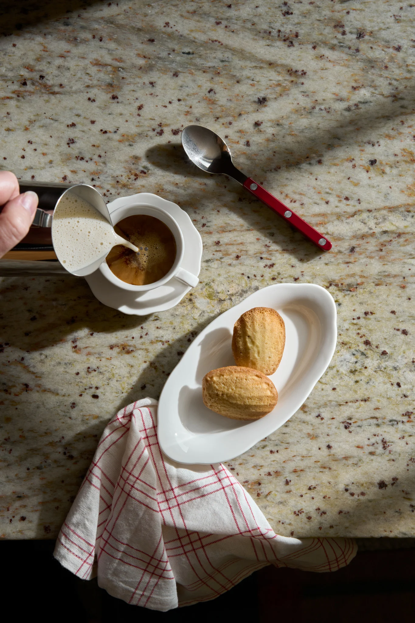Hand giet opgeschuimde melk in koffie. Twee madeleinekoekjes op een wit bord en een lepel met rood handvat rusten op een granieten aanrecht.