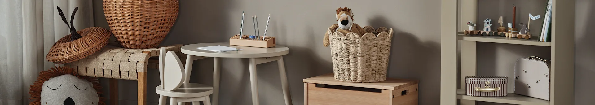 Kinderkamer met een leeuwenknuffel in een rieten mand, een kleine houten tafel met potloden en een boekenplank met speelgoed en boeken.