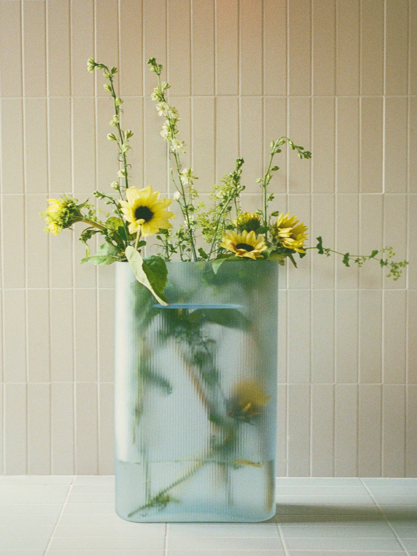 Boeket van gele zonnebloemen en groene puntige bloemen in een heldere, geribbelde rechthoekige vaas op een betegeld oppervlak.
