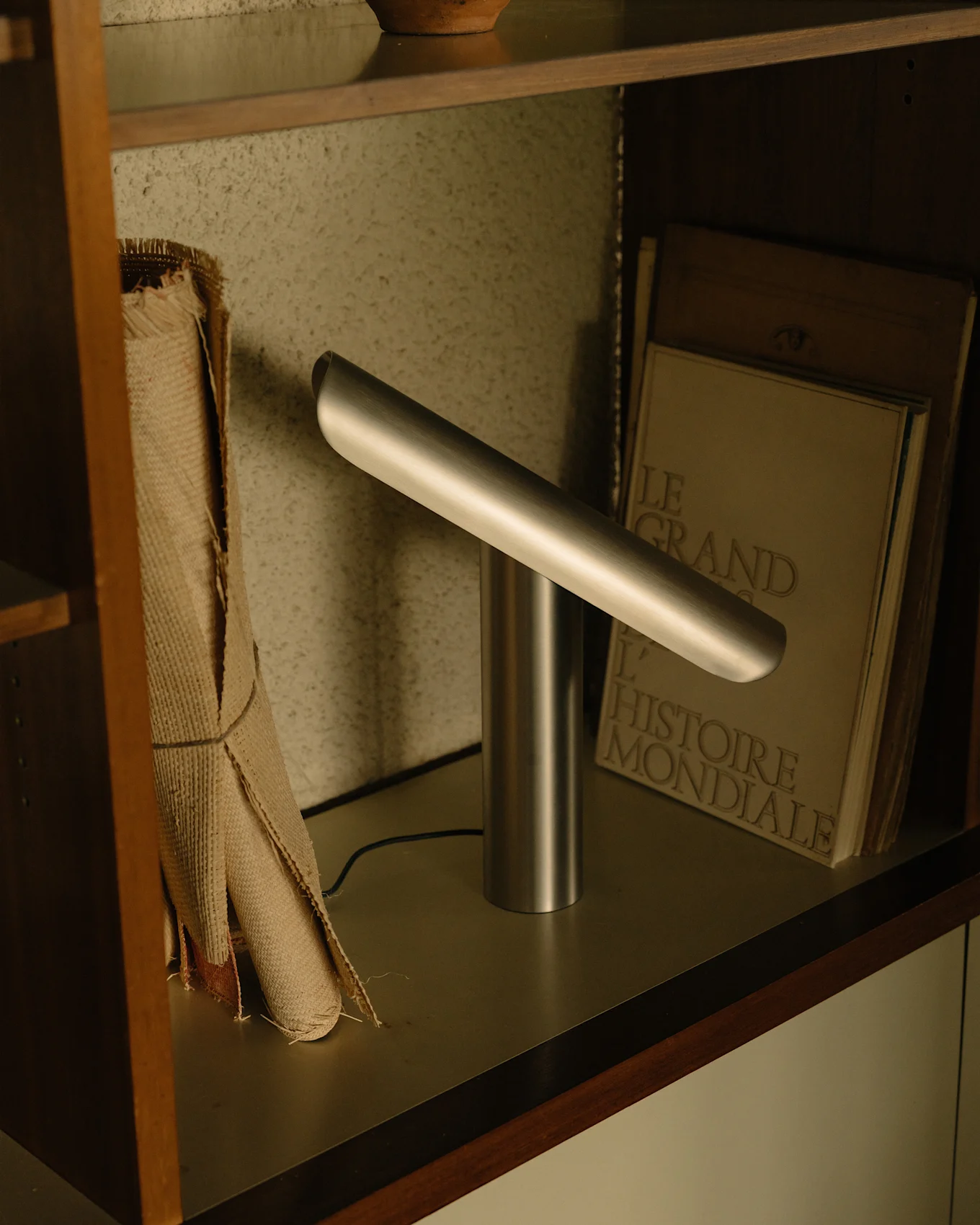 Moderne zilveren bureaulamp met een cilindrische voet en een schuin horizontale lichtbalk op een plank.