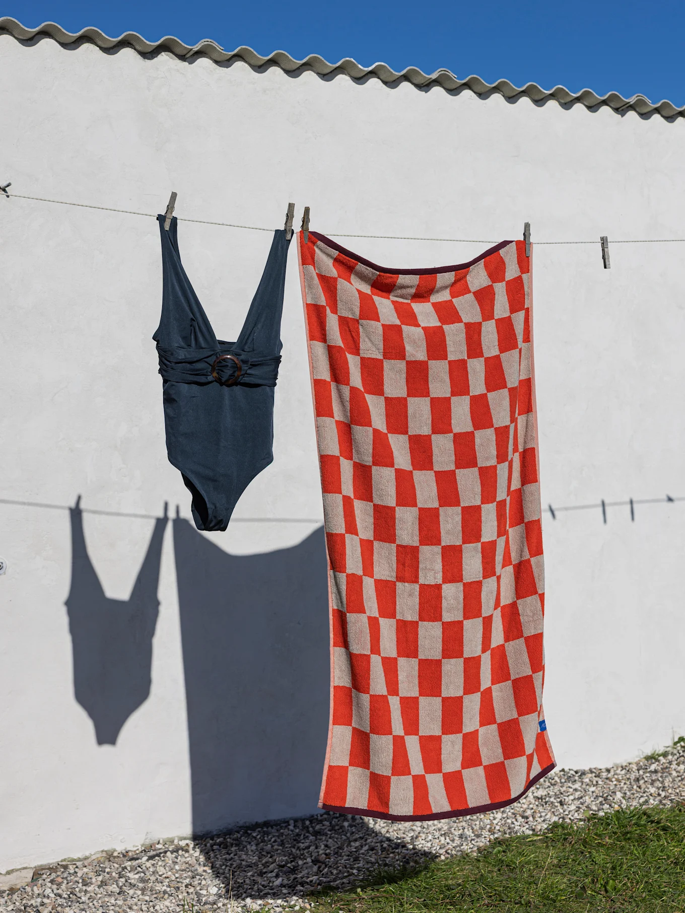 Een donkerblauwe badpak en een rood-beige geblokte strandhanddoek hangen aan een waslijn tegen een witte muur.