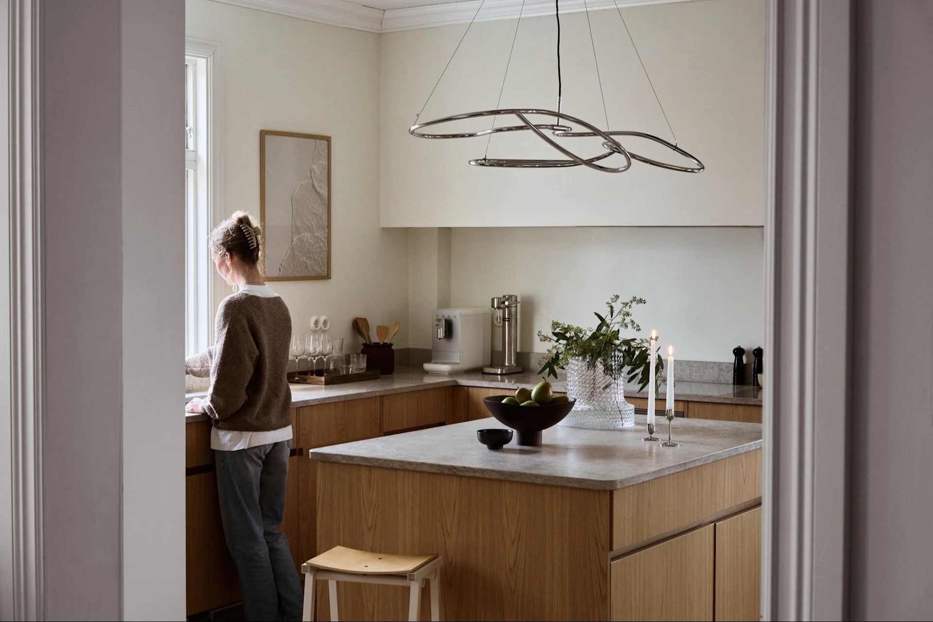 Vrouw kijkt uit een raam in een lichte, moderne keuken met lichte houten kasten, grijze werkbladen en een sculpturale hanglamp.